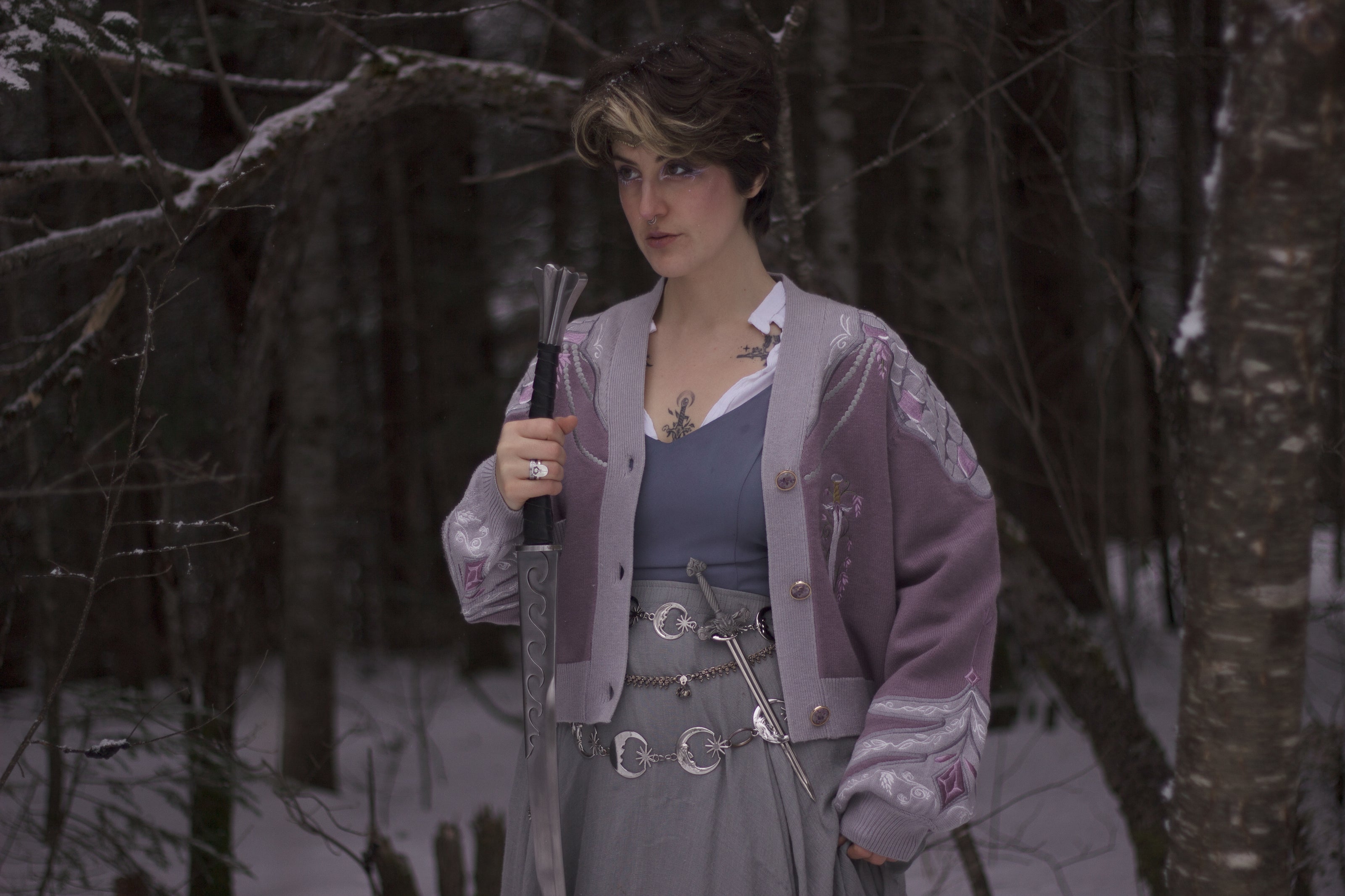 A soft button-up lavender and light plum knit cardigan shown in two styles: cropped and full-length. The design features intricate embroidery of swords and silver armour accents, evoking a medieval and fantasy-inspired look.
