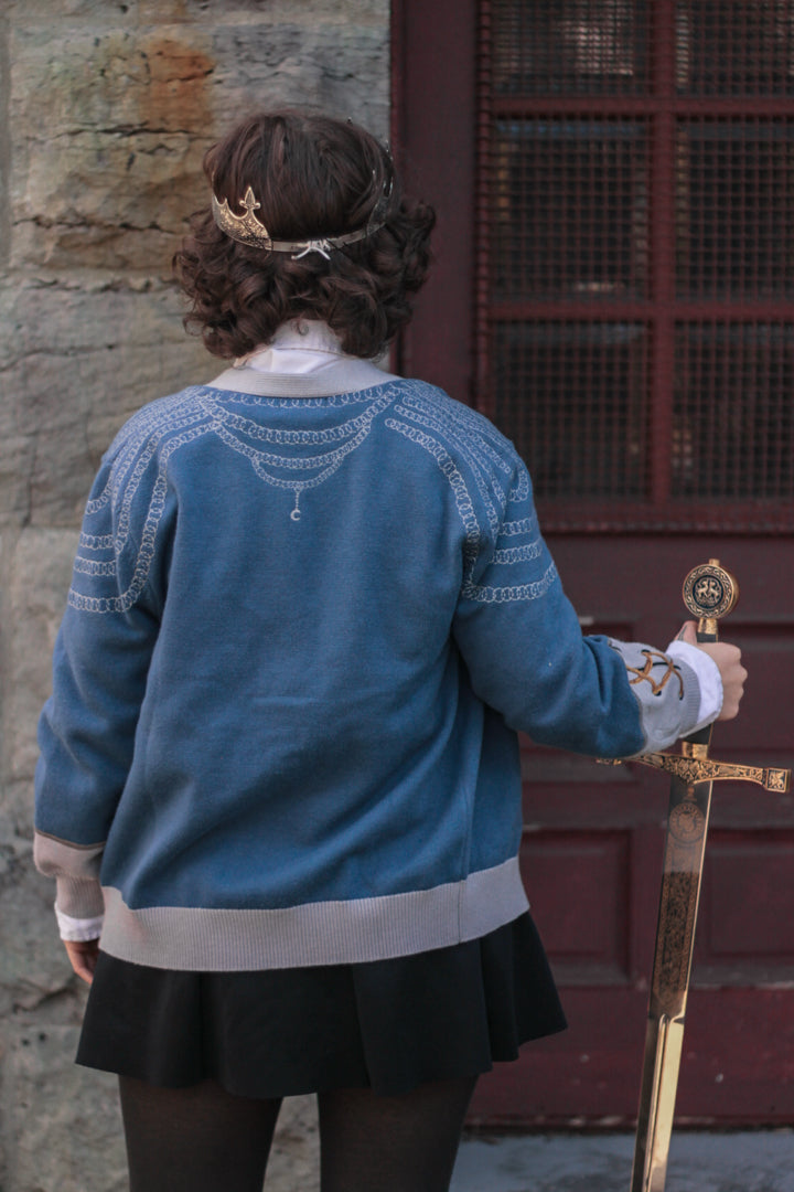 A soft, button-up, sky blue and silver knit cardigan, with black and gold moon buttons. The design features intricate embroidery of a vine wrapped sword, chainmail, and armour. Evoking a whimsical and fantasy-inspired look.