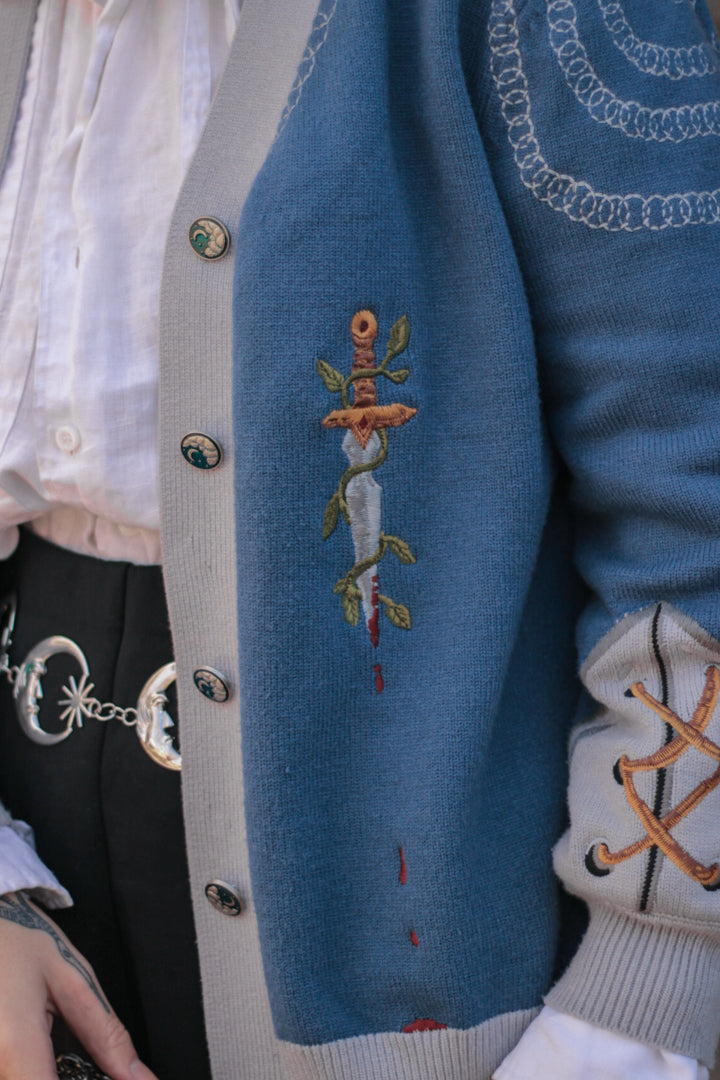A soft, button-up, sky blue and silver knit cardigan, with black and gold moon buttons. The design features intricate embroidery of a vine wrapped sword, chainmail, and armour. Evoking a whimsical and fantasy-inspired look.