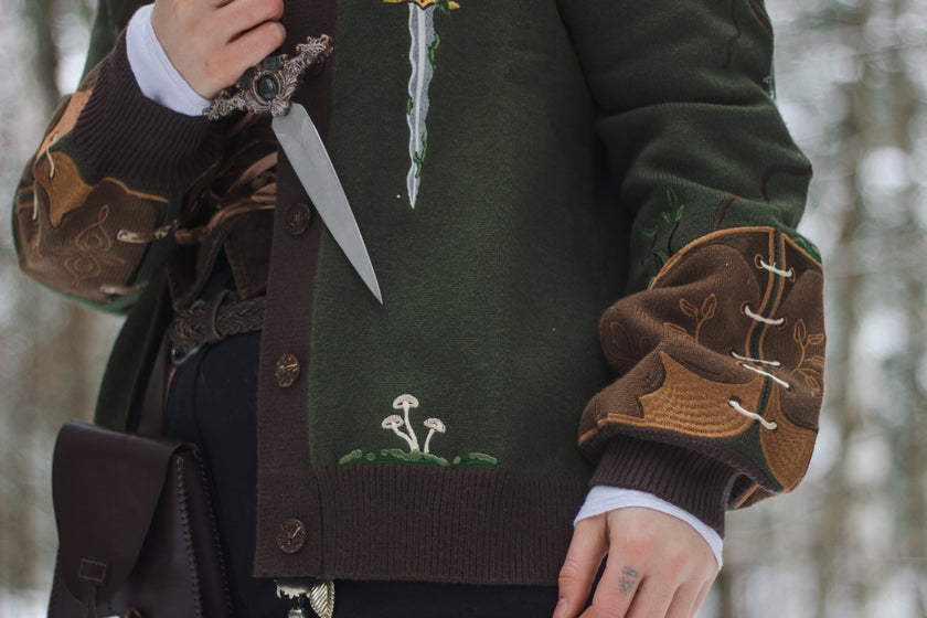  A soft button-up chestnut-brown and forest-green knit cardigan. The design features intricate embroidery of english ivy, dagger, and sturdy armour, evoking a whimsical and fantasy-inspired look.