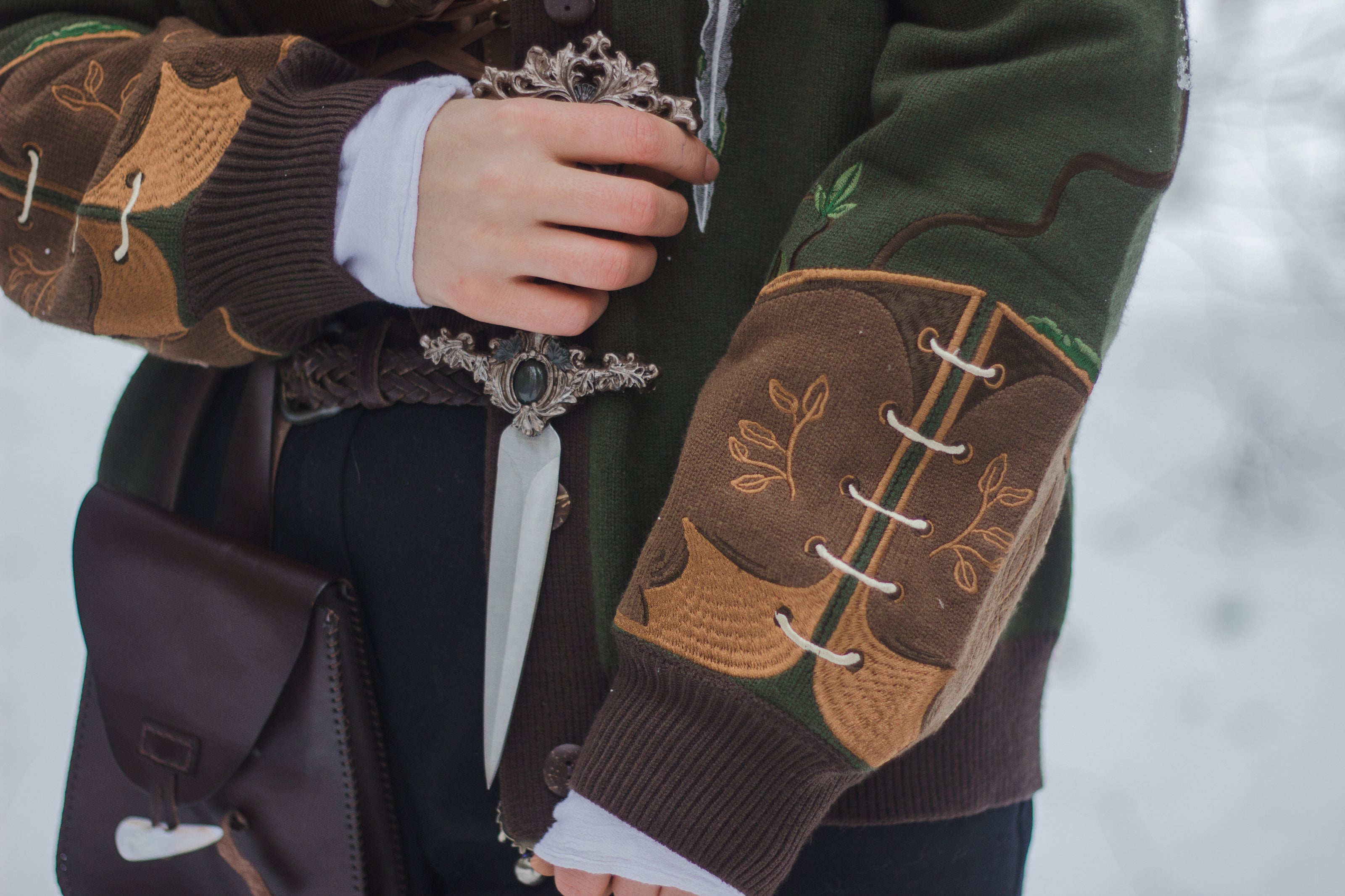  A soft button-up chestnut-brown and forest-green knit cardigan. The design features intricate embroidery of english ivy, dagger, and sturdy armour, evoking a whimsical and fantasy-inspired look.