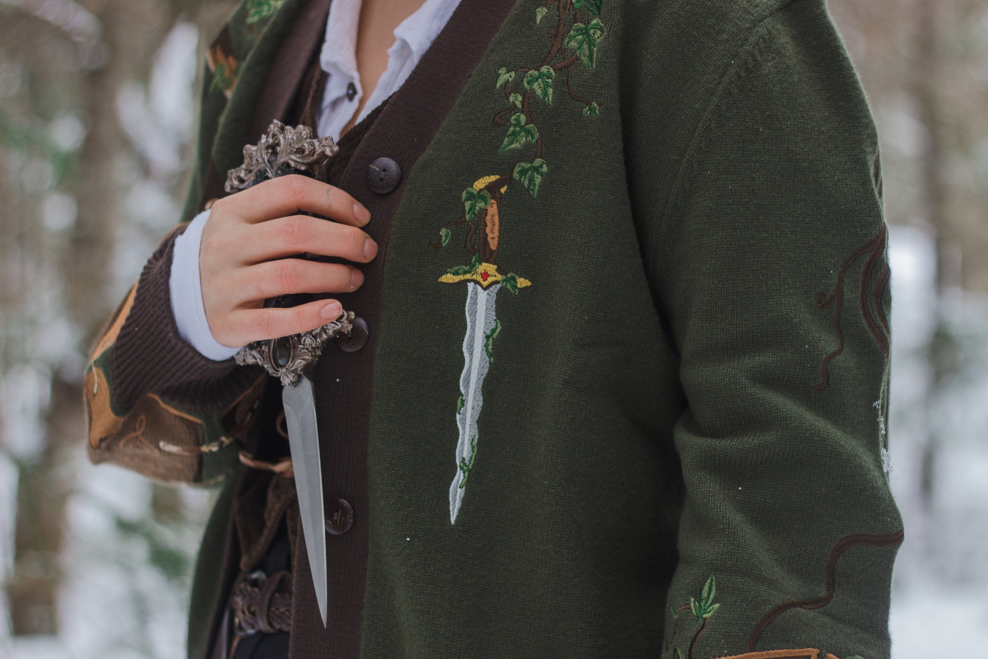  A soft button-up chestnut-brown and forest-green knit cardigan. The design features intricate embroidery of english ivy, dagger, and sturdy armour, evoking a whimsical and fantasy-inspired look.