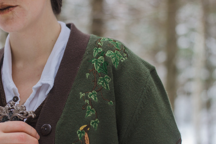  A soft button-up chestnut-brown and forest-green knit cardigan. The design features intricate embroidery of english ivy, dagger, and sturdy armour, evoking a whimsical and fantasy-inspired look.