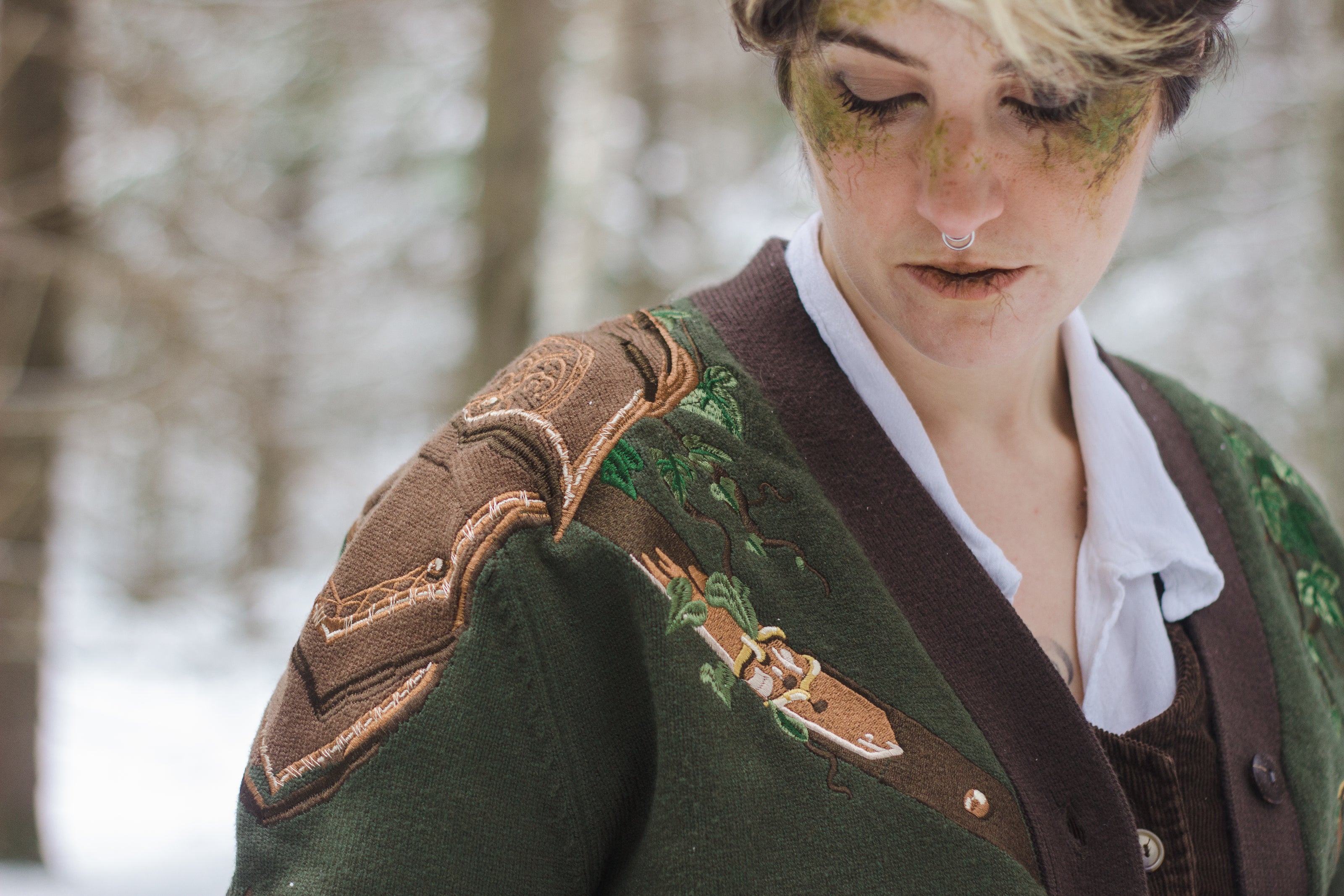  A soft button-up chestnut-brown and forest-green knit cardigan. The design features intricate embroidery of english ivy, dagger, and sturdy armour, evoking a whimsical and fantasy-inspired look.