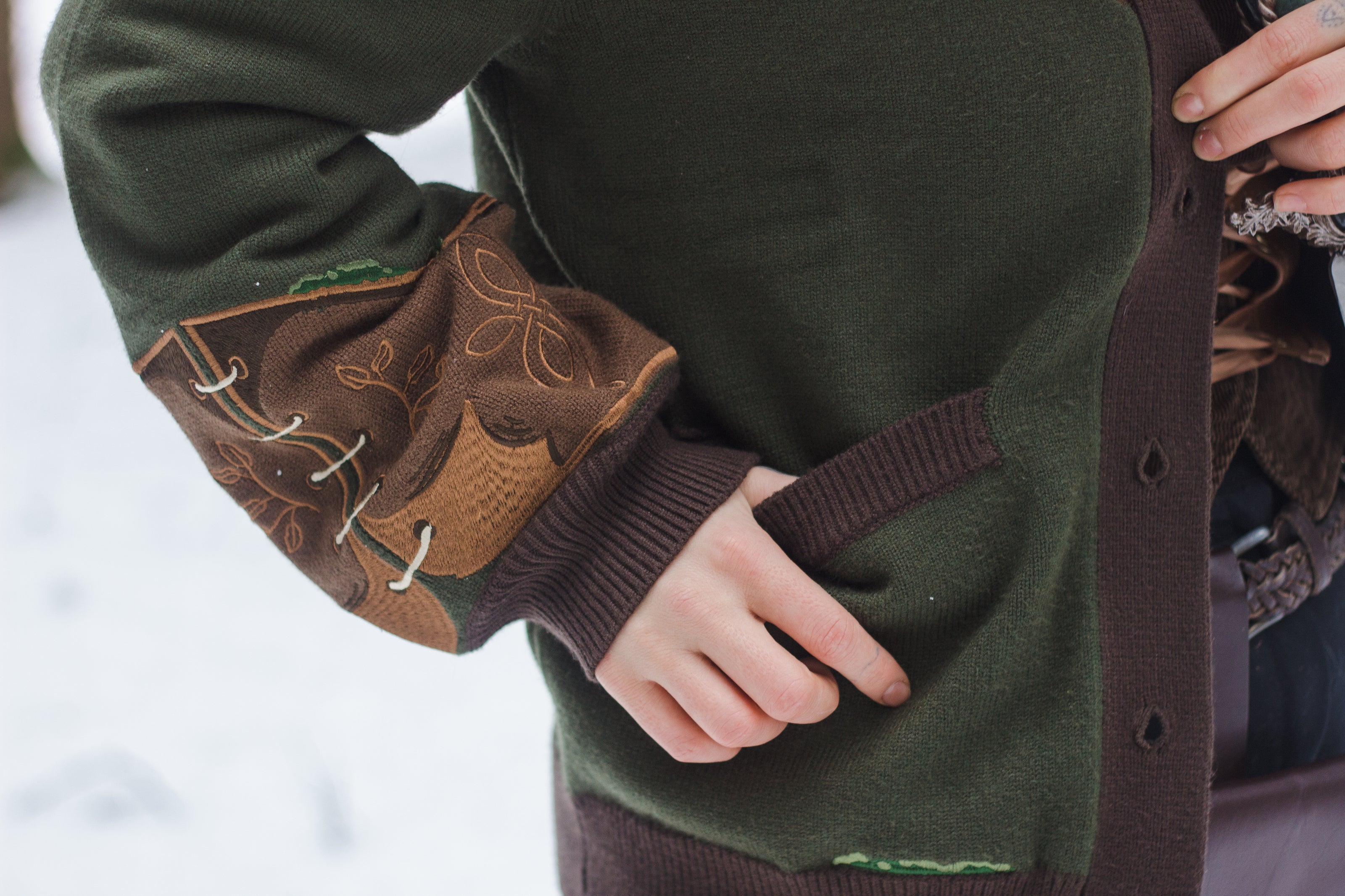  A soft button-up chestnut-brown and forest-green knit cardigan. The design features intricate embroidery of english ivy, dagger, and sturdy armour, evoking a whimsical and fantasy-inspired look.