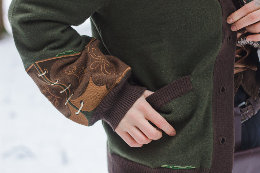  A soft button-up chestnut-brown and forest-green knit cardigan. The design features intricate embroidery of english ivy, dagger, and sturdy armour, evoking a whimsical and fantasy-inspired look.