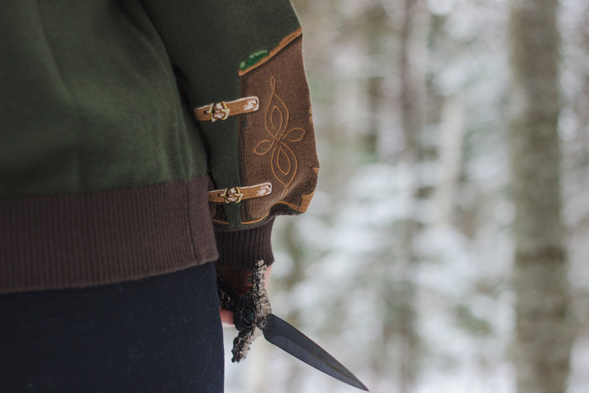  A soft button-up chestnut-brown and forest-green knit cardigan. The design features intricate embroidery of english ivy, dagger, and sturdy armour, evoking a whimsical and fantasy-inspired look.