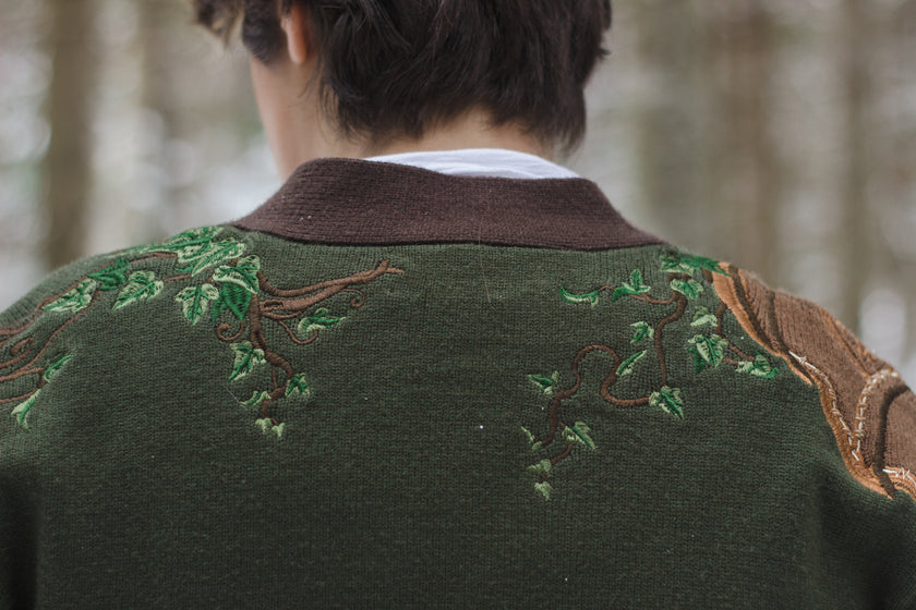  A soft button-up chestnut-brown and forest-green knit cardigan. The design features intricate embroidery of english ivy, dagger, and sturdy armour, evoking a whimsical and fantasy-inspired look.