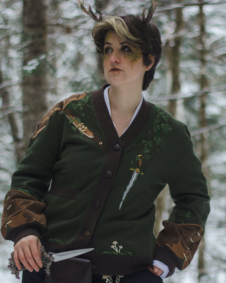  A soft button-up chestnut-brown and forest-green knit cardigan. The design features intricate embroidery of english ivy, dagger, and sturdy armour, evoking a whimsical and fantasy-inspired look.