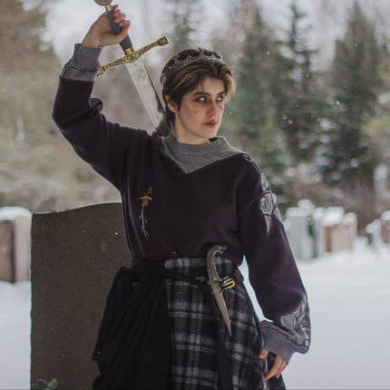 A 100% cotton, dark purple and silver knit crewneck sweater. The design features intricate embroidery of a vine wrapped sword and sturdy armour. Evoking a whimsical and fantasy-inspired look.