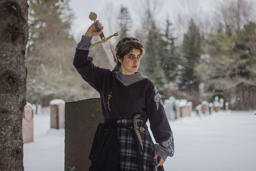 A soft, dark purple with a slight brown undertone and silver knit crewneck sweater, with red embellishments. The design features intricate embroidery of a vine wrapped sword and sturdy armour. Evoking a whimsical and fantasy-inspired look.