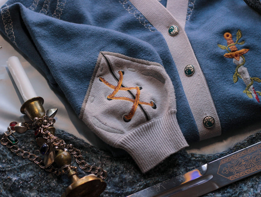 A soft, button-up, sky blue and silver knit cardigan, with black and gold moon buttons. The design features intricate embroidery of a vine wrapped sword, chainmail, and armour. Evoking a whimsical and fantasy-inspired look.