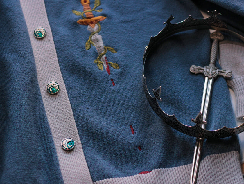 A soft, button-up, sky blue and silver knit cardigan, with black and gold moon buttons. The design features intricate embroidery of a vine wrapped sword, chainmail, and armour. Evoking a whimsical and fantasy-inspired look.
