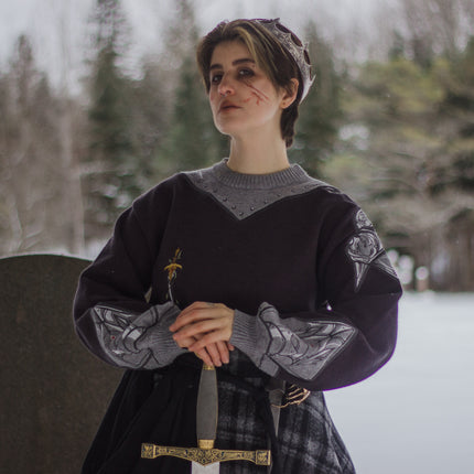 A soft, dark purple with a slight brown undertone and silver knit crewneck sweater, with red embellishments. The design features intricate embroidery of a vine wrapped sword and sturdy armour. Evoking a whimsical and fantasy-inspired look.