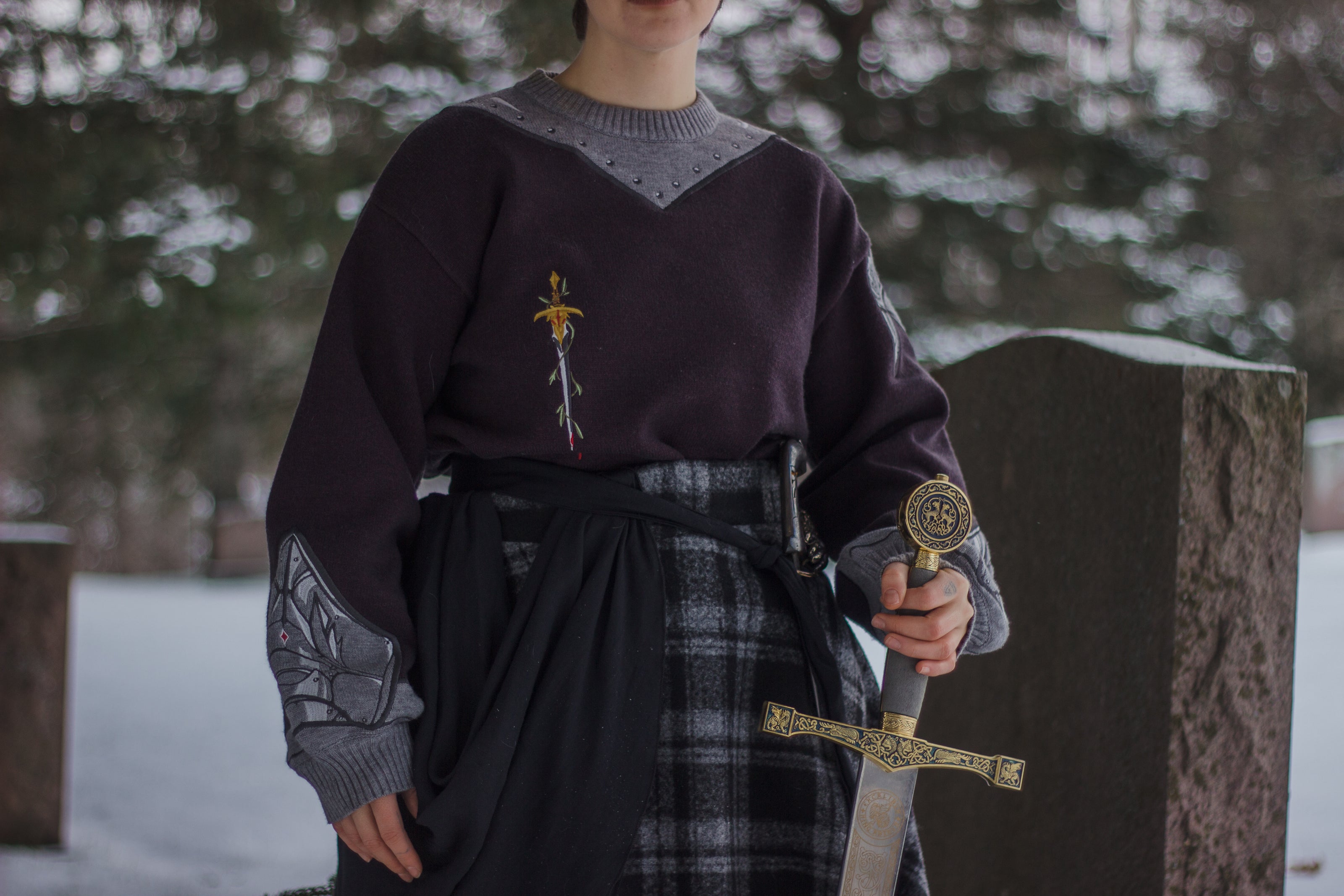 A soft, dark purple with a slight brown undertone and silver knit crewneck sweater, with red embellishments. The design features intricate embroidery of a vine wrapped sword and sturdy armour. Evoking a whimsical and fantasy-inspired look.