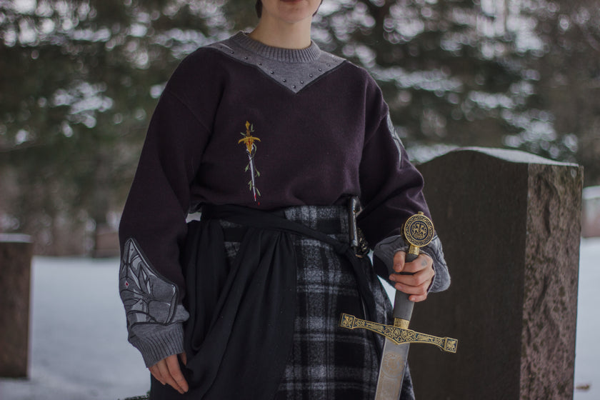 A soft, dark purple with a slight brown undertone and silver knit crewneck sweater, with red embellishments. The design features intricate embroidery of a vine wrapped sword and sturdy armour. Evoking a whimsical and fantasy-inspired look.