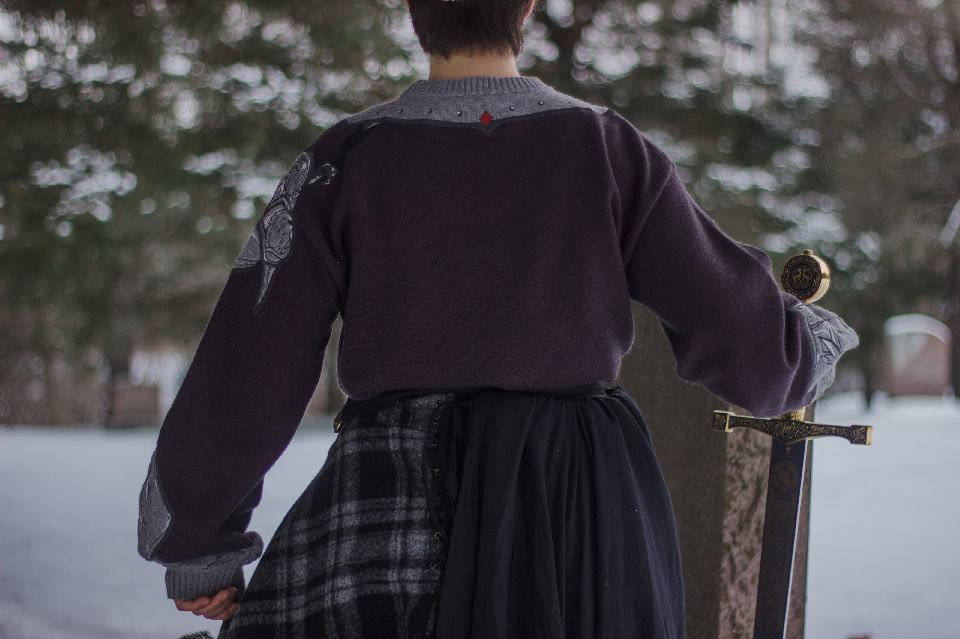 A soft, dark purple with a slight brown undertone and silver knit crewneck sweater, with red embellishments. The design features intricate embroidery of a vine wrapped sword and sturdy armour. Evoking a whimsical and fantasy-inspired look.