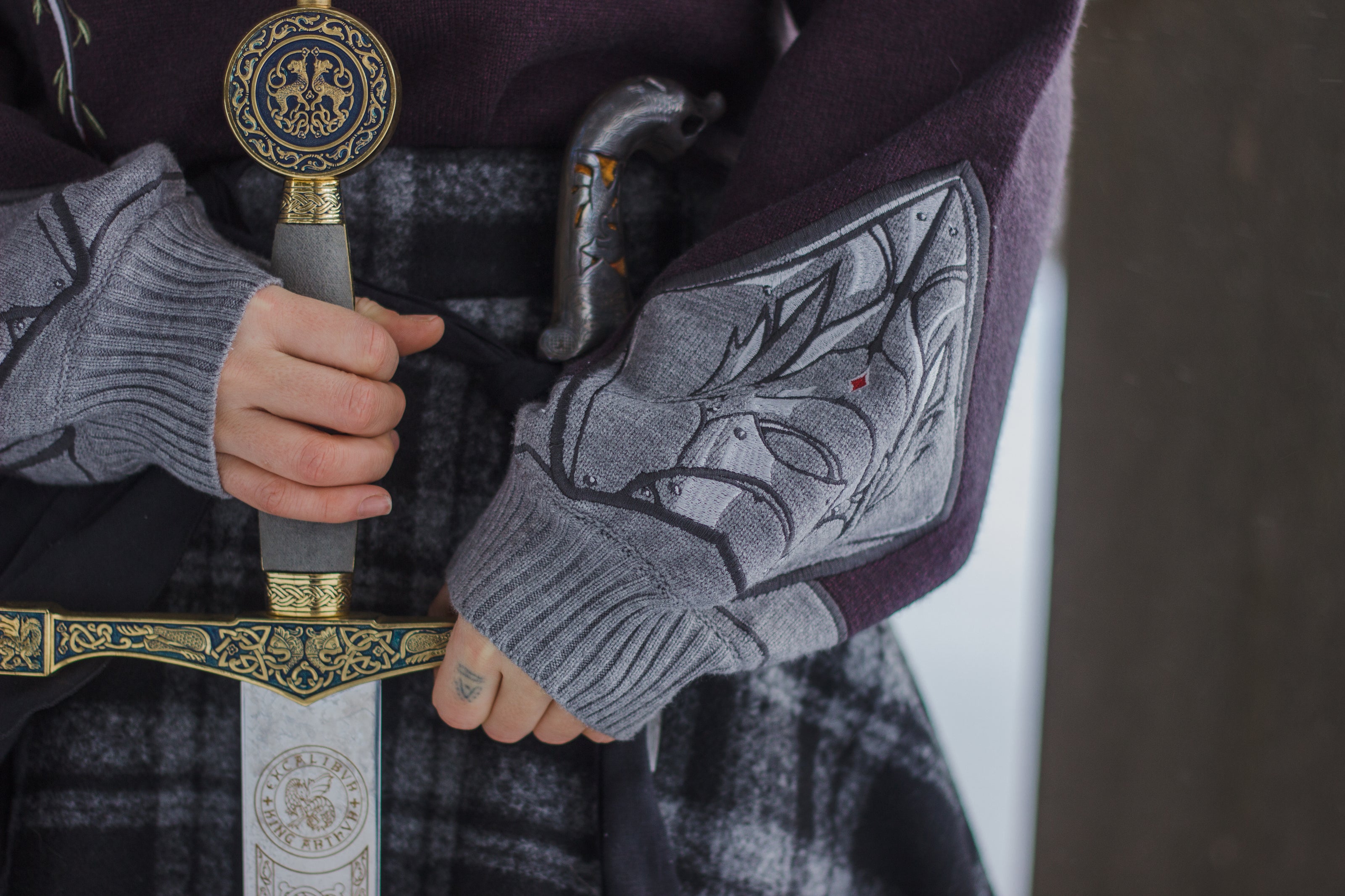 A soft, dark purple with a slight brown undertone and silver knit crewneck sweater, with red embellishments. The design features intricate embroidery of a vine wrapped sword and sturdy armour. Evoking a whimsical and fantasy-inspired look.