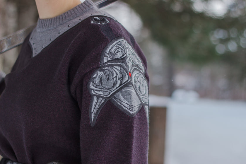 A soft, dark purple with a slight brown undertone and silver knit crewneck sweater, with red embellishments. The design features intricate embroidery of a vine wrapped sword and sturdy armour. Evoking a whimsical and fantasy-inspired look.