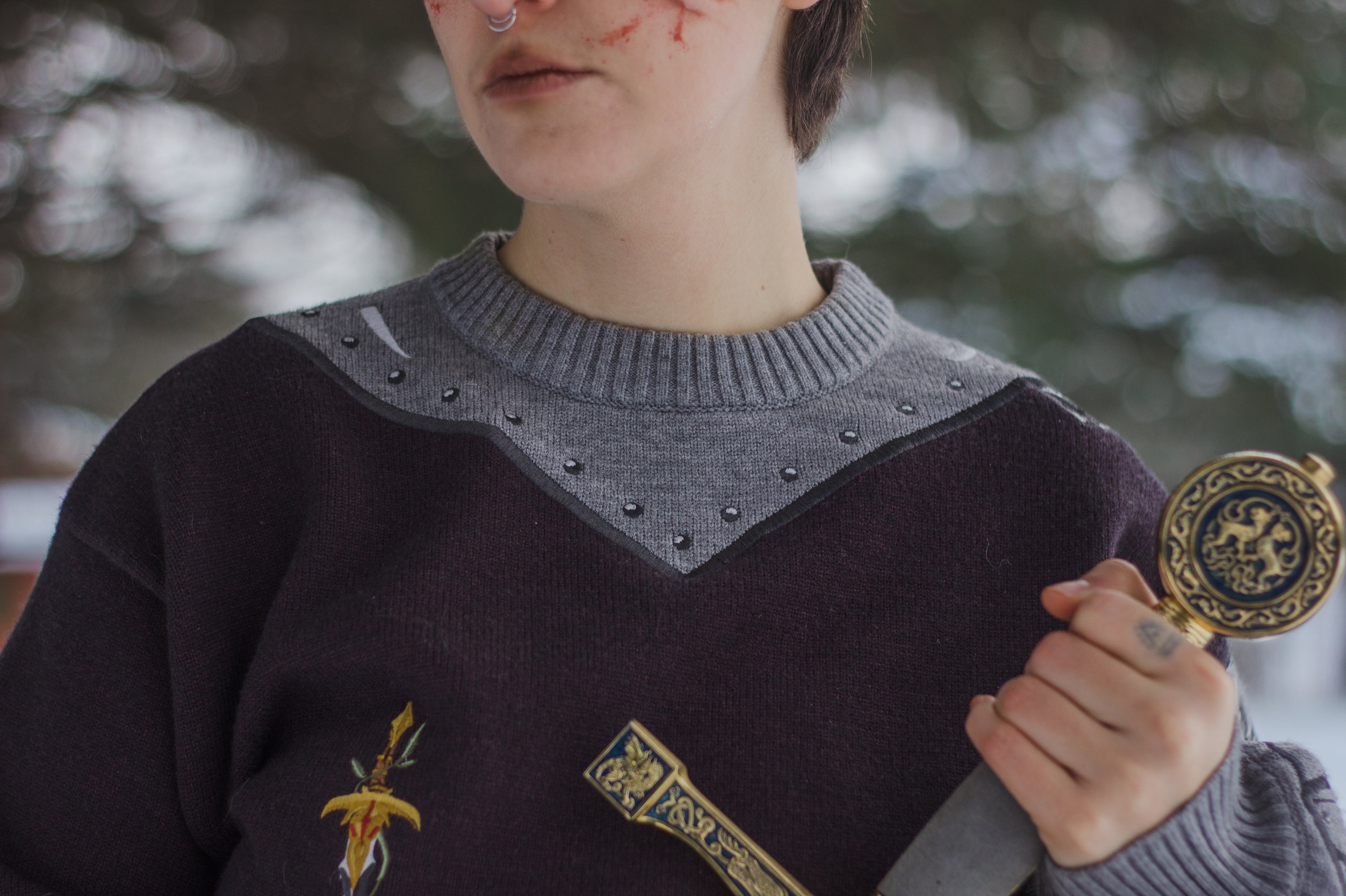 A soft, dark purple with a slight brown undertone and silver knit crewneck sweater, with red embellishments. The design features intricate embroidery of a vine wrapped sword and sturdy armour. Evoking a whimsical and fantasy-inspired look.
