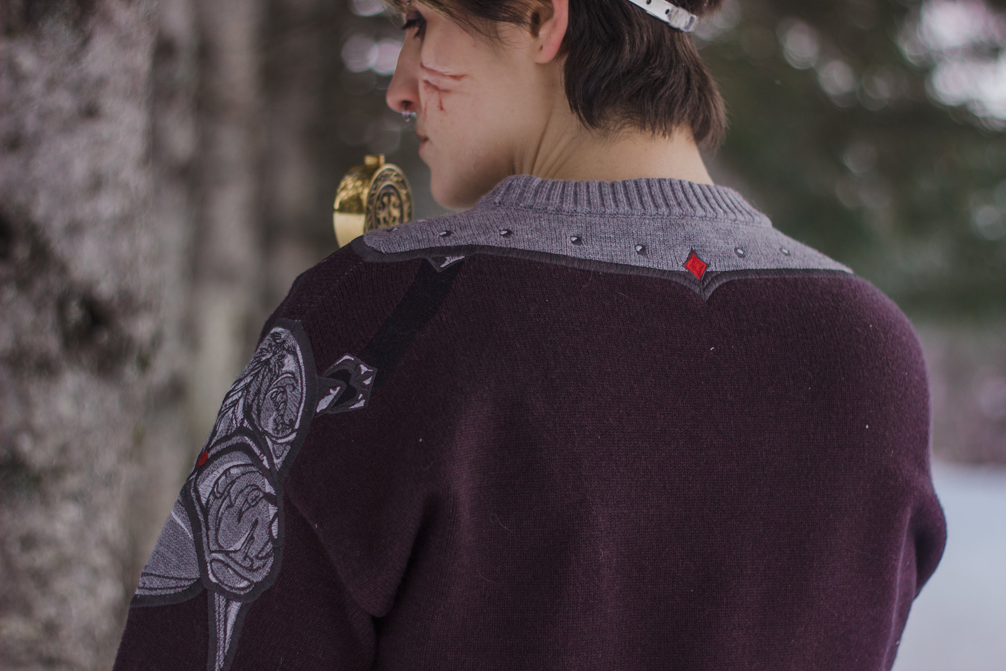 A soft, dark purple with a slight brown undertone and silver knit crewneck sweater, with red embellishments. The design features intricate embroidery of a vine wrapped sword and sturdy armour. Evoking a whimsical and fantasy-inspired look.