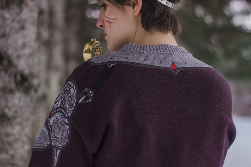 A soft, dark purple with a slight brown undertone and silver knit crewneck sweater, with red embellishments. The design features intricate embroidery of a vine wrapped sword and sturdy armour. Evoking a whimsical and fantasy-inspired look.