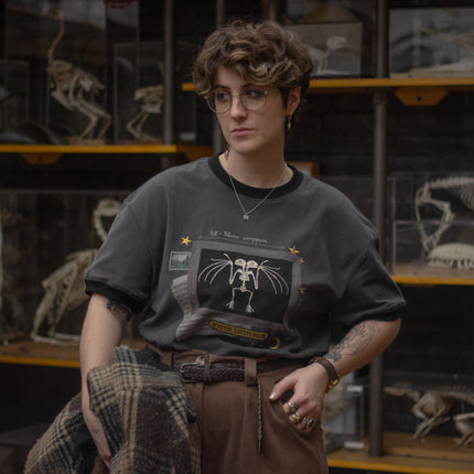 Charcoal gray cotton T-shirt featuring an embroidered skeletal illustration of Myotis lucifugus (a little brown bat) framed within a vintage-style display. The design includes handwritten text noting bat wingspan, golden star and moon accents, and a collage of vintage-inspired elements.