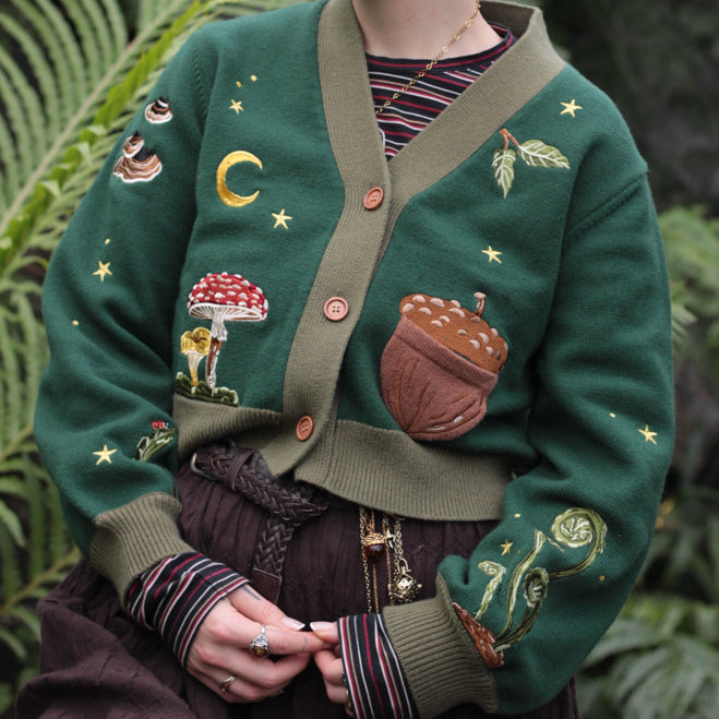 A soft, knit, forest-green cardigan with intricate woodland embroidery. The cardigan features details of a vibrant red toadstool mushroom, a large textured acorn pocket, golden stars, a crescent moon, fiddle heads, turkey-tail mushrooms, and leafy vines on the sleeves and chest. Available in both cropped and full-length styles.