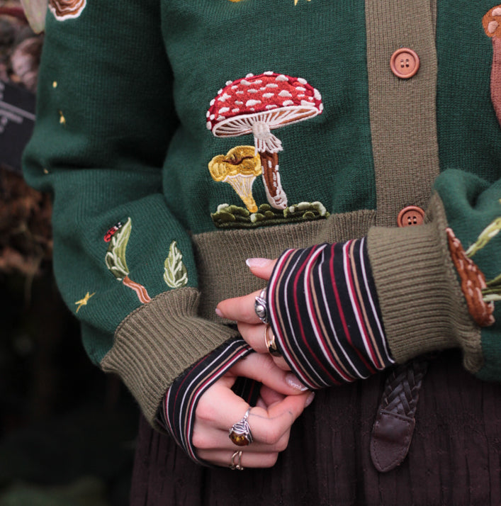 A soft, knit, forest-green cardigan with intricate woodland embroidery. The cardigan features details of a vibrant red toadstool mushroom, a large textured acorn pocket, golden stars, a crescent moon, fiddle heads, turkey-tail mushrooms, and leafy vines on the sleeves and chest. Available in both cropped and full-length styles.
