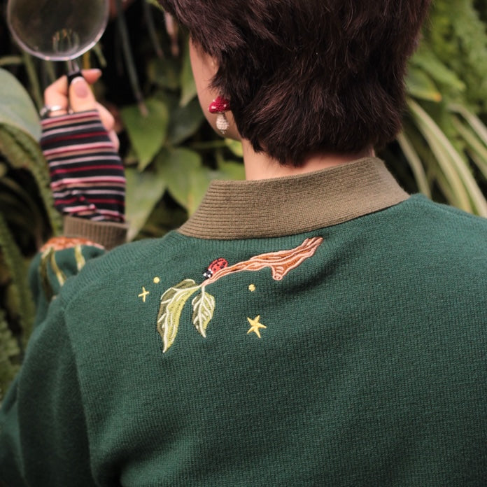 A soft, knit, forest-green cardigan with intricate woodland embroidery. The cardigan features details of a vibrant red toadstool mushroom, a large textured acorn pocket, golden stars, a crescent moon, fiddle heads, turkey-tail mushrooms, and leafy vines on the sleeves and chest. Available in both cropped and full-length styles.