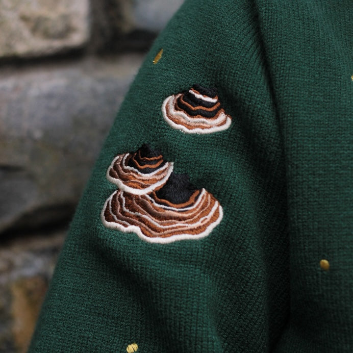 A soft, knit, forest-green cardigan with intricate woodland embroidery. The cardigan features details of a vibrant red toadstool mushroom, a large textured acorn pocket, golden stars, a crescent moon, fiddle heads, turkey-tail mushrooms, and leafy vines on the sleeves and chest. Available in both cropped and full-length styles.
