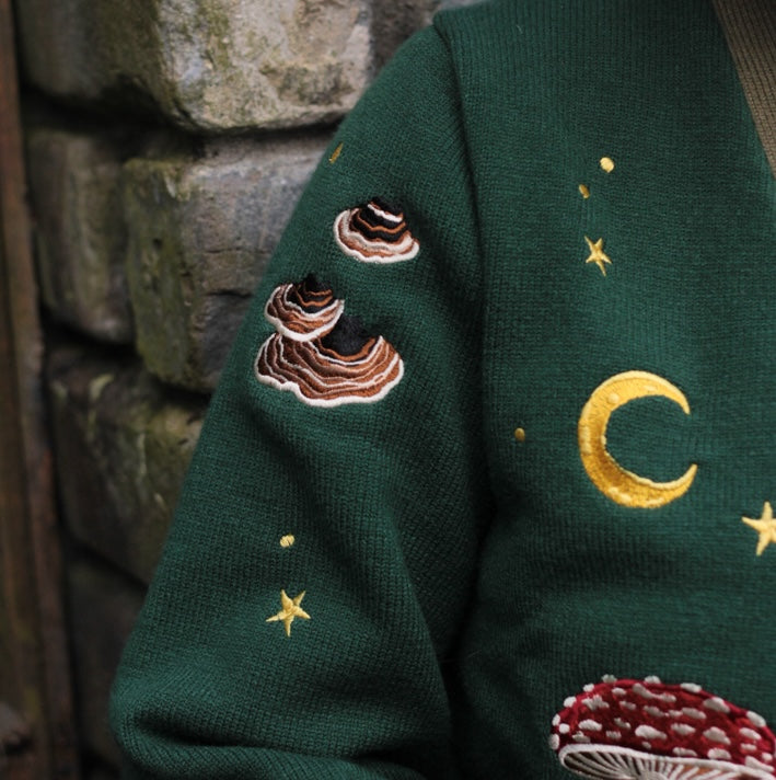 A soft, knit, forest-green cardigan with intricate woodland embroidery. The cardigan features details of a vibrant red toadstool mushroom, a large textured acorn pocket, golden stars, a crescent moon, fiddle heads, turkey-tail mushrooms, and leafy vines on the sleeves and chest. Available in both cropped and full-length styles.