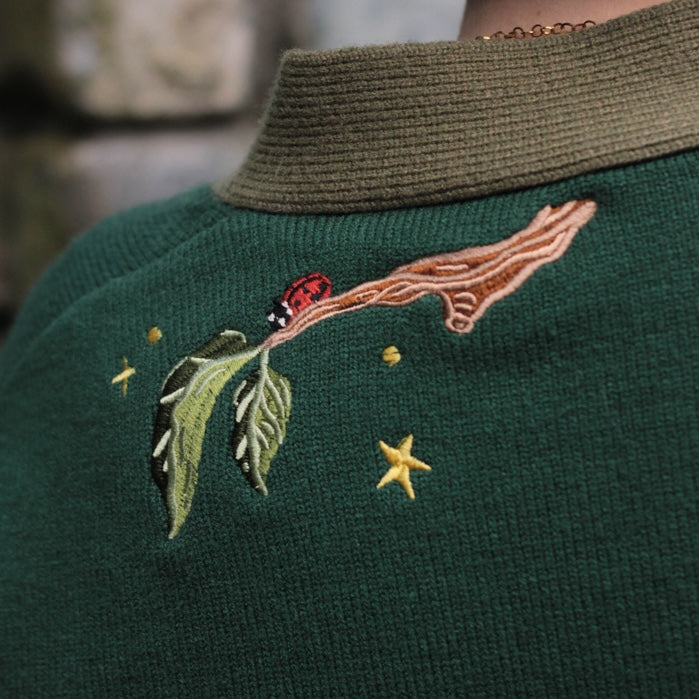 A soft, knit, forest-green cardigan with intricate woodland embroidery. The cardigan features details of a vibrant red toadstool mushroom, a large textured acorn pocket, golden stars, a crescent moon, fiddle heads, turkey-tail mushrooms, and leafy vines on the sleeves and chest. Available in both cropped and full-length styles.