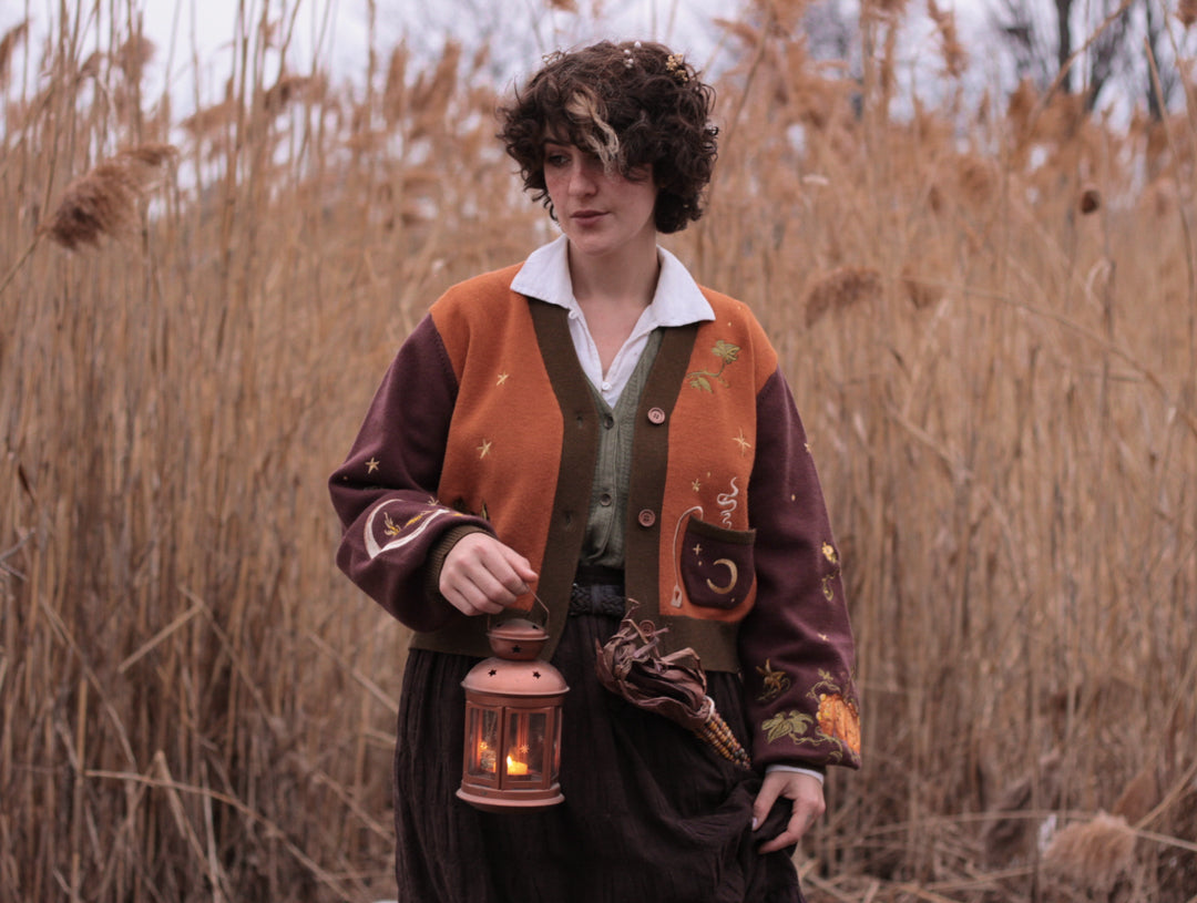 A soft, knit, pumpkin-orange and mulberry cardigan with intricate autumn themed embroidery. The cardigan features details of a ripe pumpkin, a large scythe, golden stars, a crescent moon, and leafy vines on the sleeves and chest. Available in both cropped and full-length styles.