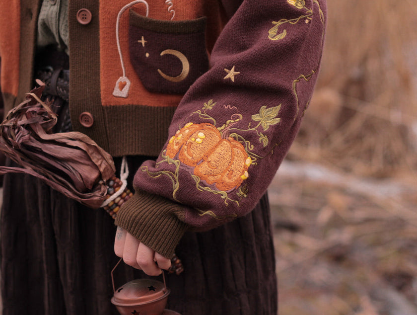 A soft, knit, pumpkin-orange and mulberry cardigan with intricate autumn themed embroidery. The cardigan features details of a ripe pumpkin, a large scythe, golden stars, a crescent moon, and leafy vines on the sleeves and chest. Available in both cropped and full-length styles.