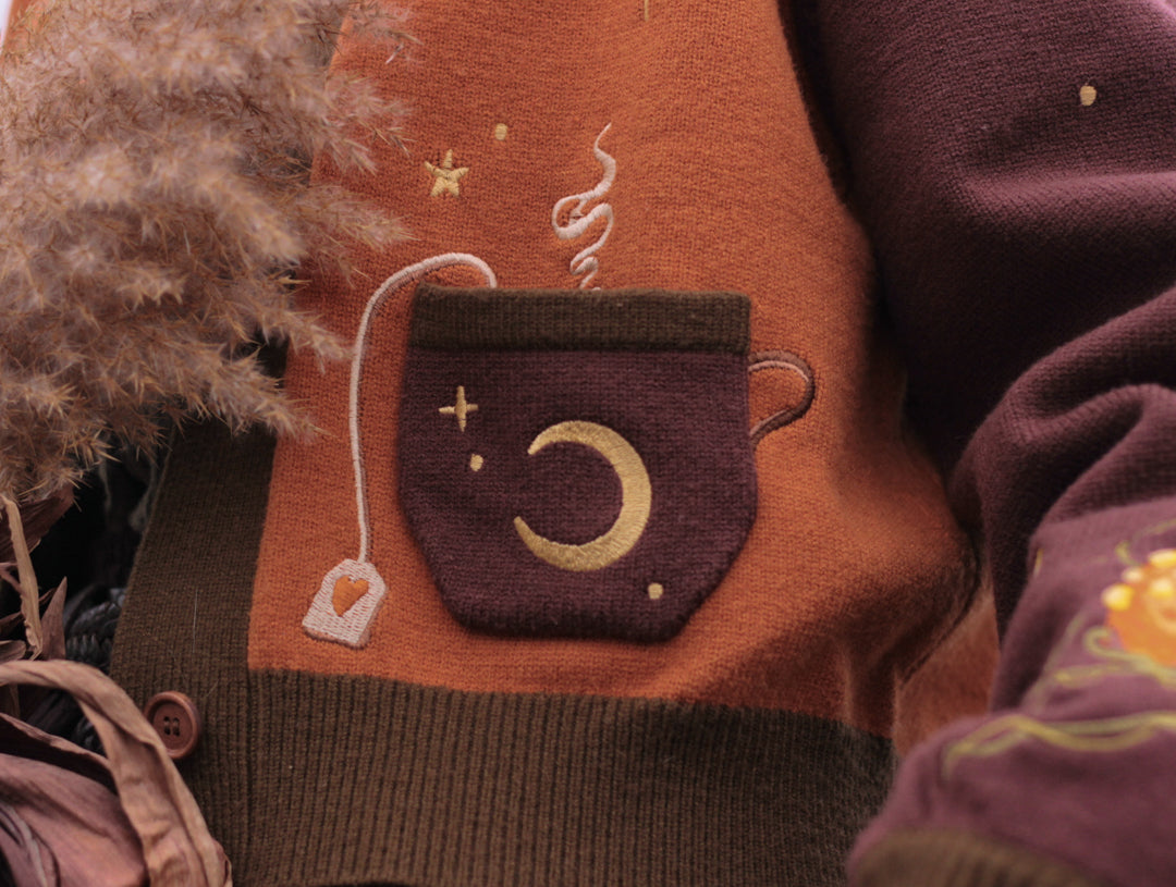 A soft, knit, pumpkin-orange and mulberry cardigan with intricate autumn themed embroidery. The cardigan features details of a ripe pumpkin, a large scythe, golden stars, a crescent moon, and leafy vines on the sleeves and chest. Available in both cropped and full-length styles.