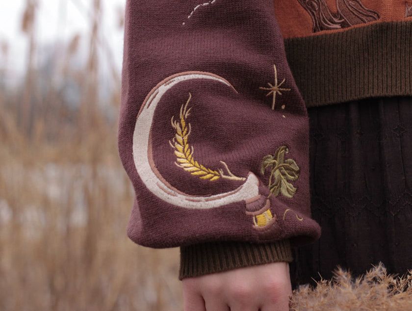 A soft, knit, pumpkin-orange and mulberry cardigan with intricate autumn themed embroidery. The cardigan features details of a ripe pumpkin, a large scythe, golden stars, a crescent moon, and leafy vines on the sleeves and chest. Available in both cropped and full-length styles.