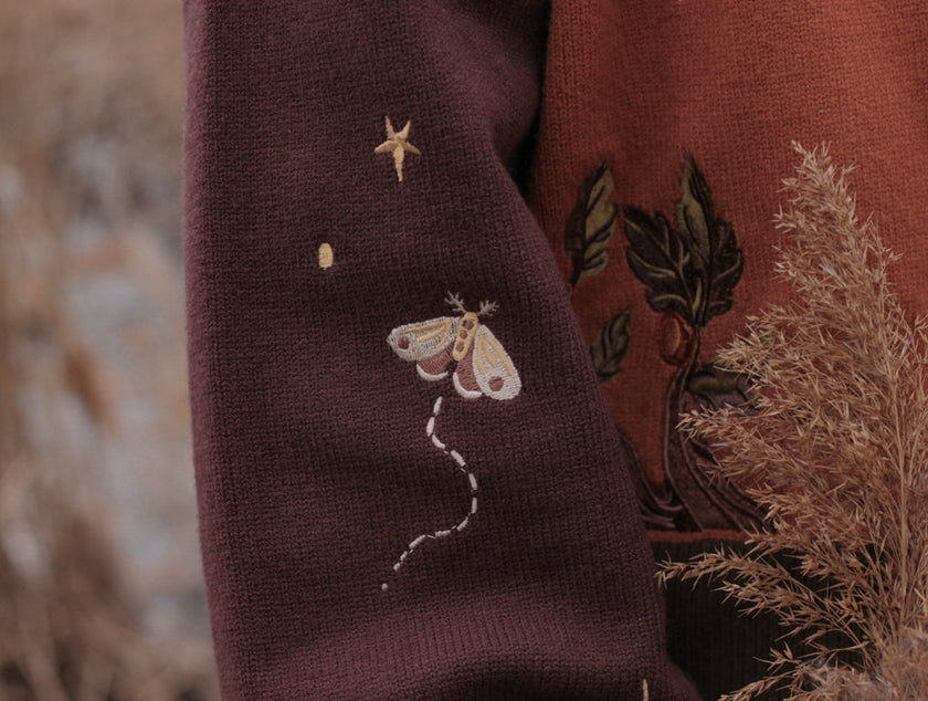 A soft, knit, pumpkin-orange and mulberry cardigan with intricate autumn themed embroidery. The cardigan features details of a ripe pumpkin, a large scythe, golden stars, a crescent moon, and leafy vines on the sleeves and chest. Available in both cropped and full-length styles.