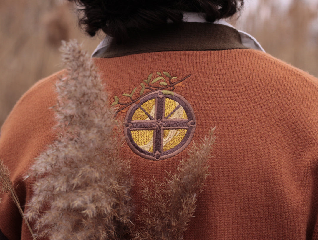 A soft, knit, pumpkin-orange and mulberry cardigan with intricate autumn themed embroidery. The cardigan features details of a ripe pumpkin, a large scythe, golden stars, a crescent moon, and leafy vines on the sleeves and chest. Available in both cropped and full-length styles.