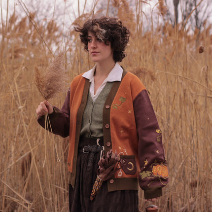 A soft, knit, pumpkin-orange and mulberry cardigan with intricate autumn themed embroidery. The cardigan features details of a ripe pumpkin, a large scythe, golden stars, a crescent moon, and leafy vines on the sleeves and chest. Available in both cropped and full-length styles.