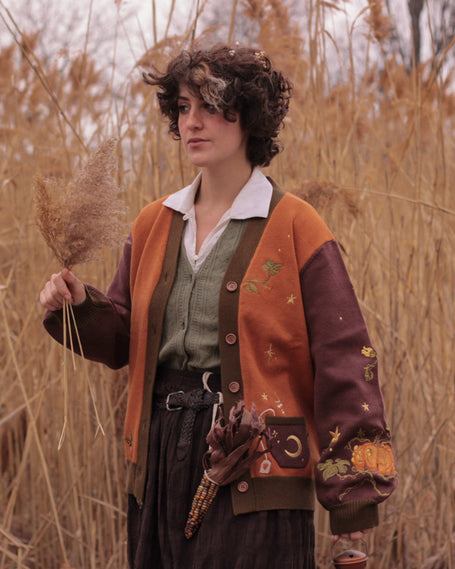A soft, knit, pumpkin-orange and mulberry cardigan with intricate autumn themed embroidery. The cardigan features details of a ripe pumpkin, a large scythe, golden stars, a crescent moon, and leafy vines on the sleeves and chest. Available in both cropped and full-length styles.
