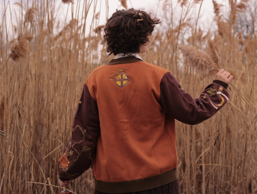 A soft, knit, pumpkin-orange and mulberry cardigan with intricate autumn themed embroidery. The cardigan features details of a ripe pumpkin, a large scythe, golden stars, a crescent moon, and leafy vines on the sleeves and chest. Available in both cropped and full-length styles.