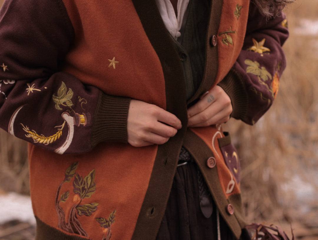A soft, knit, pumpkin-orange and mulberry cardigan with intricate autumn themed embroidery. The cardigan features details of a ripe pumpkin, a large scythe, golden stars, a crescent moon, and leafy vines on the sleeves and chest. Available in both cropped and full-length styles.