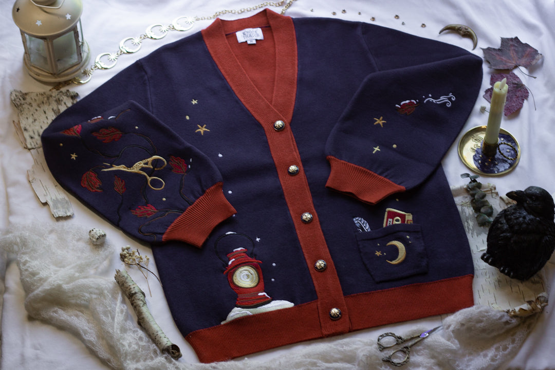 A soft, button-up, oversized, knit cardigan, in shades of navy blue, crimson red, and vintage gold. The cardigan features whimsical embroidered details of autumn leaves, moon and stars, a snowy lantern, and a bluebirds feather.