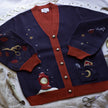 A soft, button-up, oversized, knit cardigan, in shades of navy blue, crimson red, and vintage gold. The cardigan features whimsical embroidered details of autumn leaves, moon and stars, a snowy lantern, and a bluebirds feather.