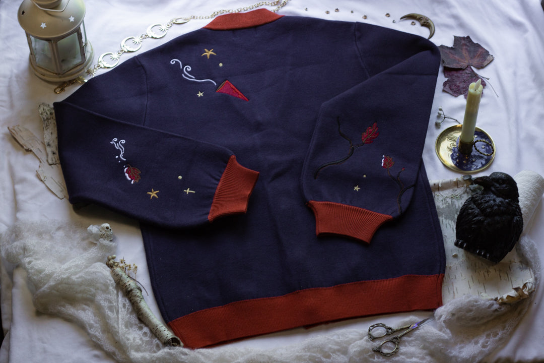 A soft, button-up, oversized, knit cardigan, in shades of navy blue, crimson red, and vintage gold. The cardigan features whimsical embroidered details of autumn leaves, moon and stars, a snowy lantern, and a bluebirds feather.