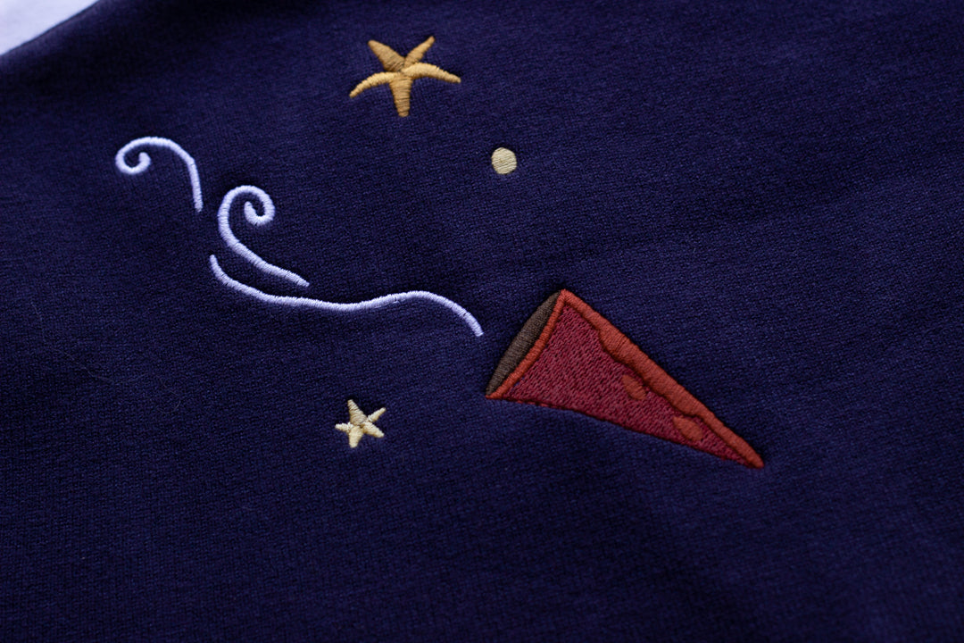 A soft, button-up, oversized, knit cardigan, in shades of navy blue, crimson red, and vintage gold. The cardigan features whimsical embroidered details of autumn leaves, moon and stars, a snowy lantern, and a bluebirds feather.