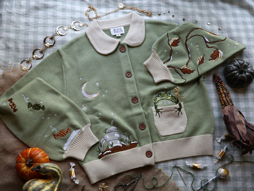 A soft, button-up, cropped, knit cardigan, in shades of soft sage and warm cream. The cardigan features whimsical embroidered details of autumn leaves, candy, snow covered branches, teapot, and froggy friend.