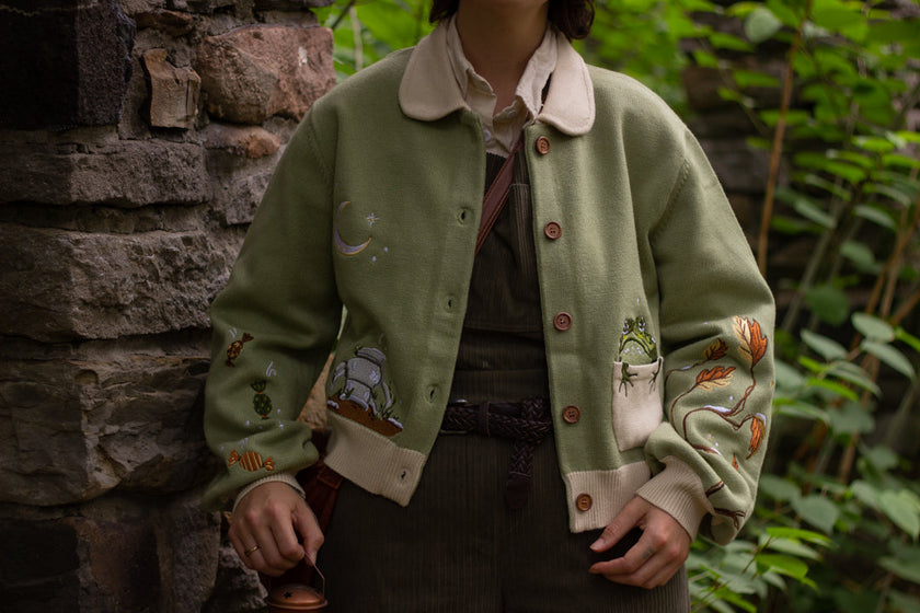 A soft, button-up, cropped, knit cardigan, in shades of soft sage and warm cream. The cardigan features whimsical embroidered details of autumn leaves, candy, snow covered branches, teapot, and froggy friend.