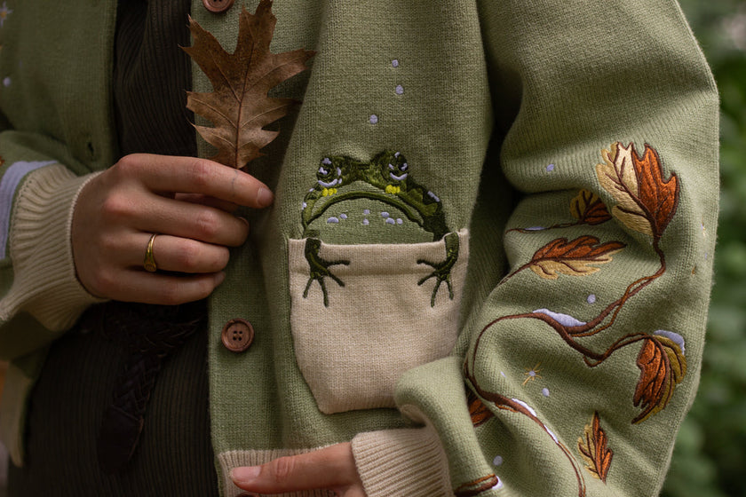 A soft, button-up, cropped, knit cardigan, in shades of soft sage and warm cream. The cardigan features whimsical embroidered details of autumn leaves, candy, snow covered branches, teapot, and froggy friend.